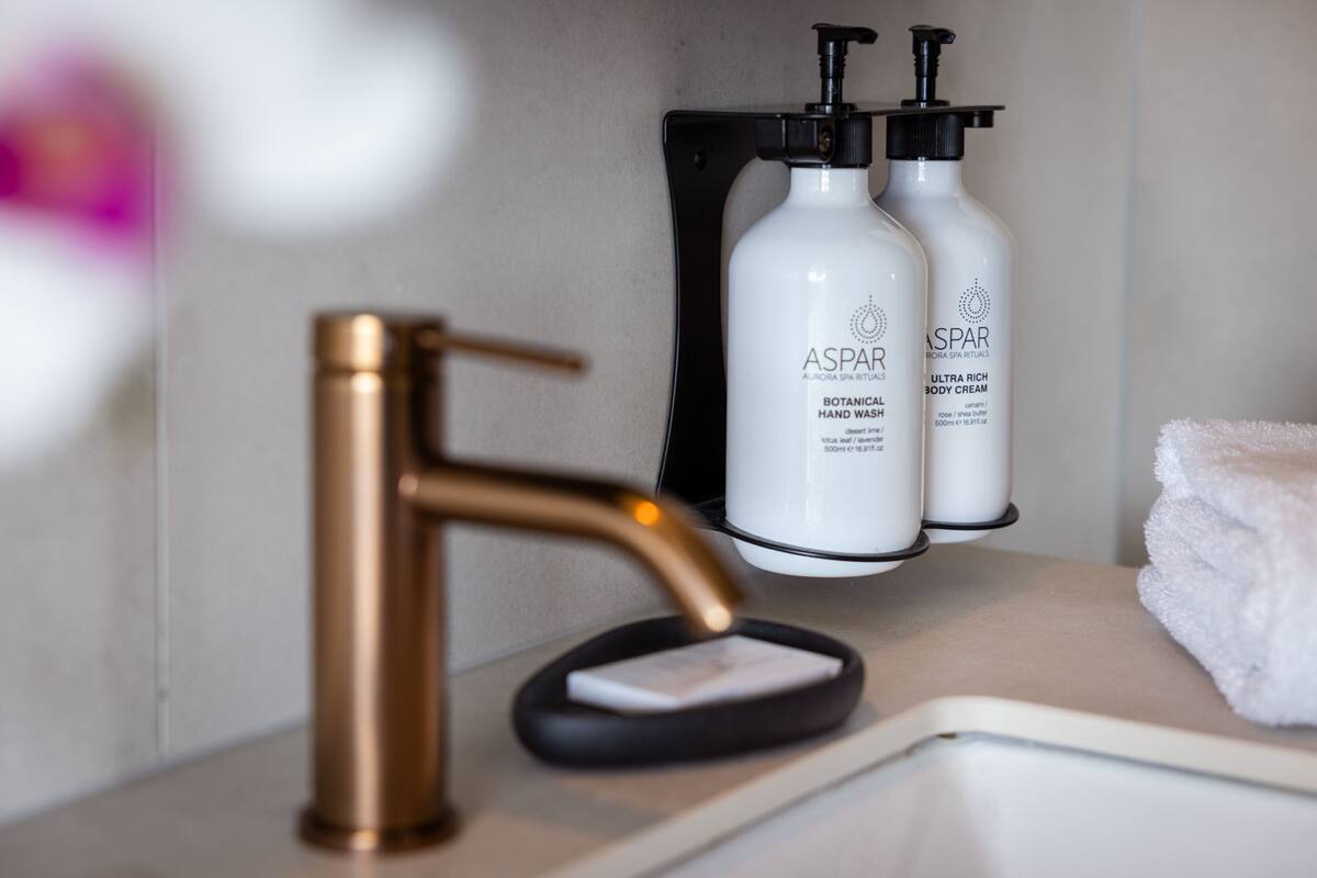 Een badkamer met een gouden kraan, een zwarte zeepbak en twee witte flessen met de aanduiding 'ASPAR' op een beige aanrecht.
