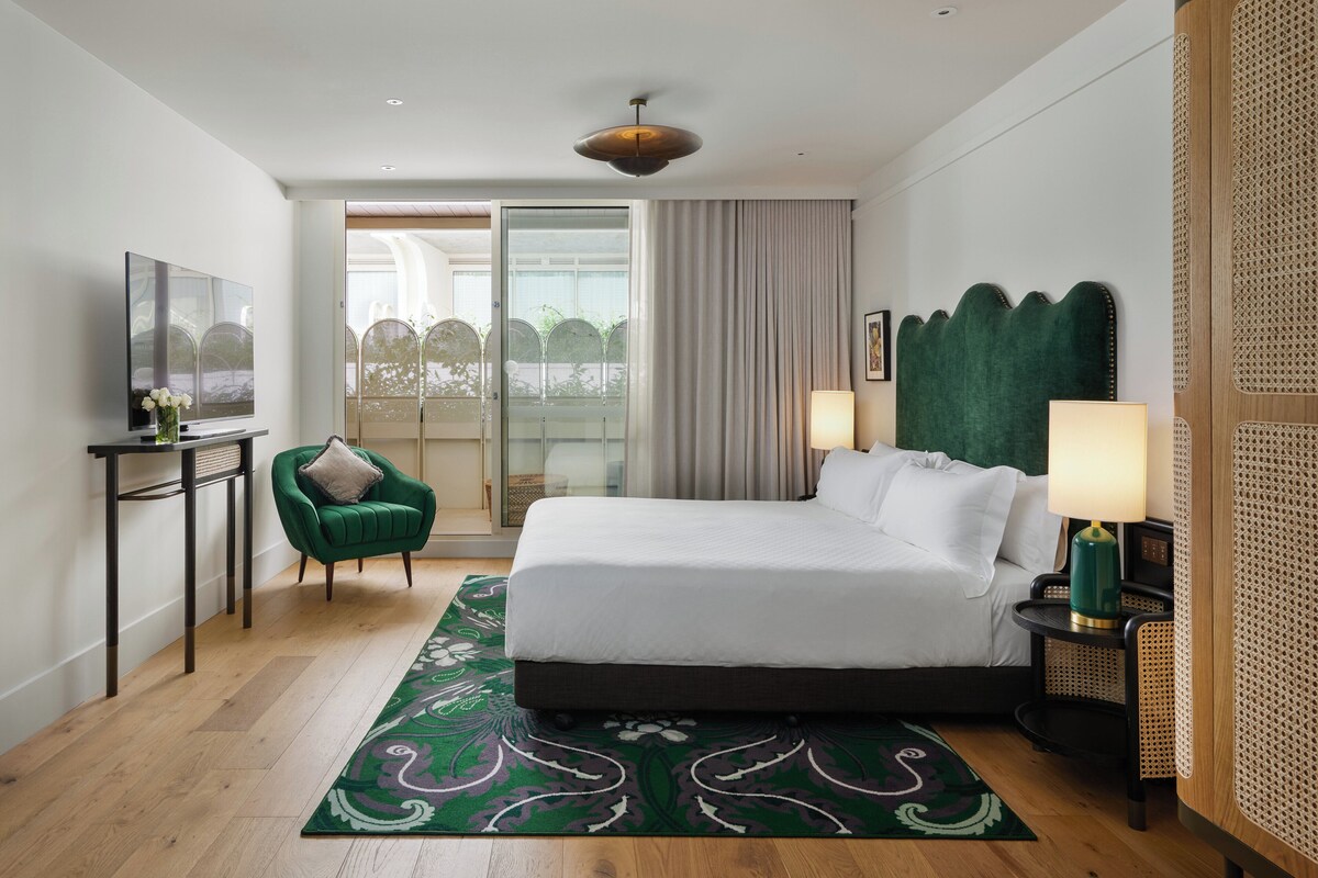 A modern hotel room with a large bed, a green armchair, a flat-screen TV, and a balcony with a view.