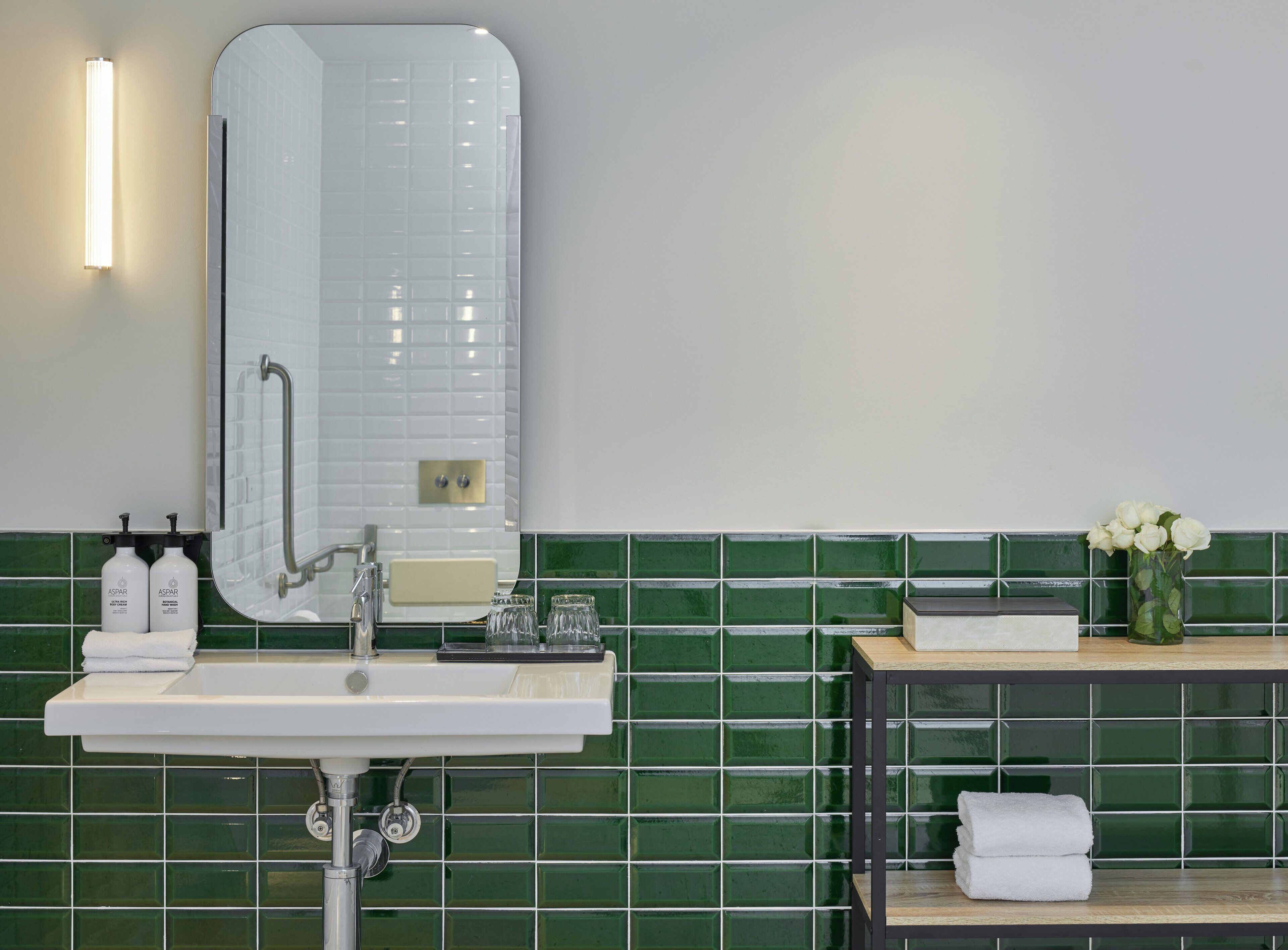A bathroom with a white sink, a mirror, green tiled walls, and a small table with a vase of flowers.
