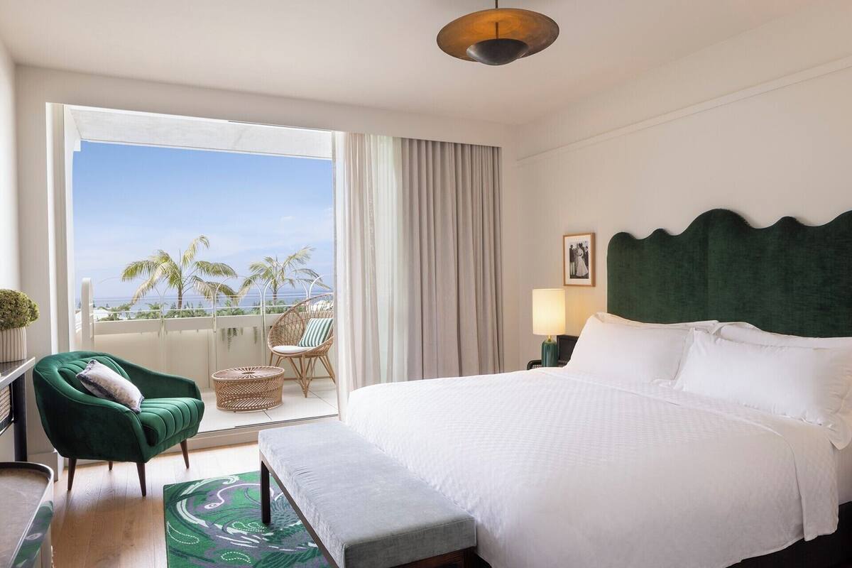 Een hotelkamer met een bed, een stoel, een klein tafeltje, een lamp en uitzicht op palmbomen.