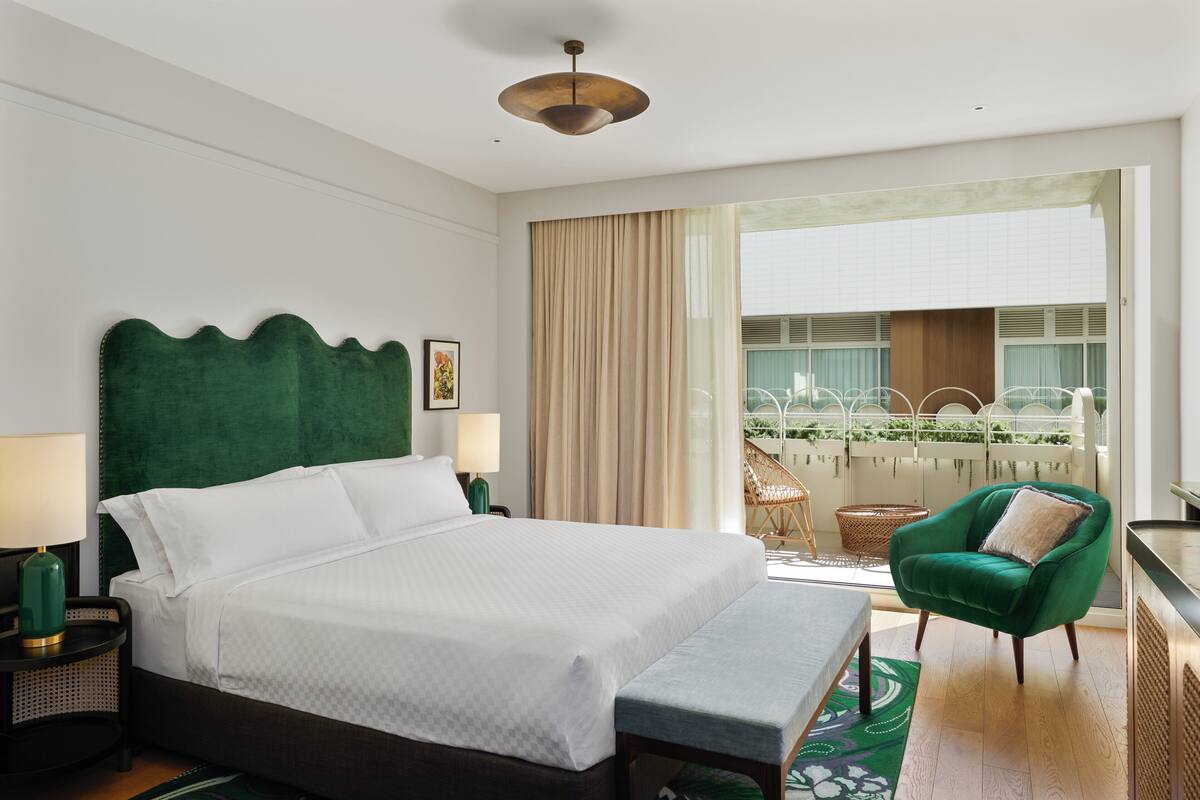 Een hotelkamer met een groot bed, een groene fauteuil, een balkon met uitzicht op groen en een plafondlamp.