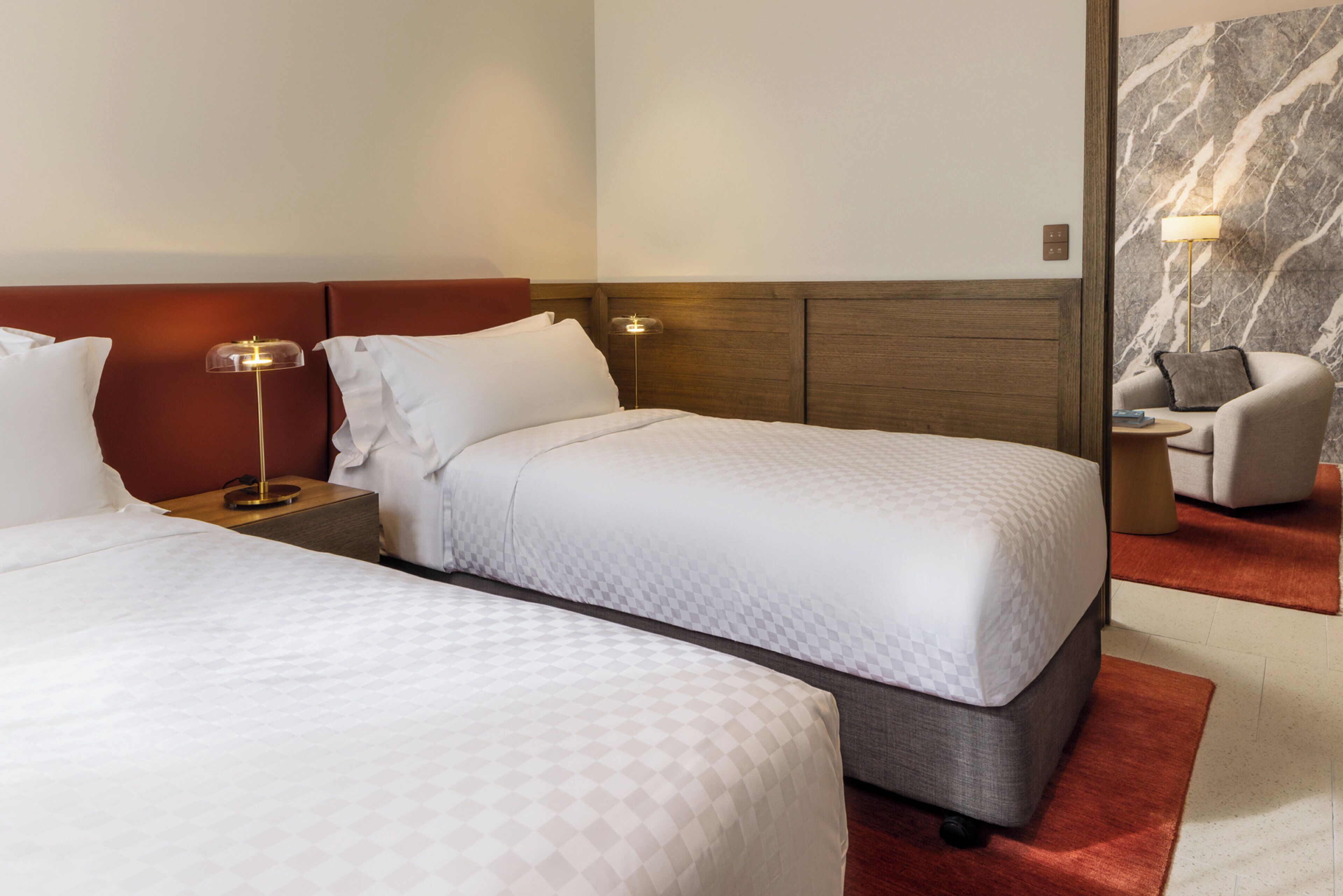 Een hotelkamer met twee bedden, een nachtkastje met lamp, een stoel en een marmeren muur.