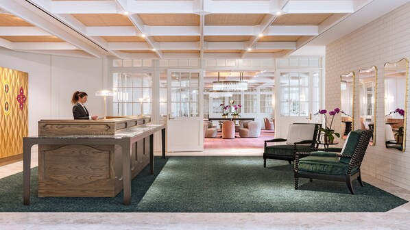 Zona de recepción con un mostrador de madera, alfombra verde y un área de asientos con una butaca tapizada de verde y una mesa.