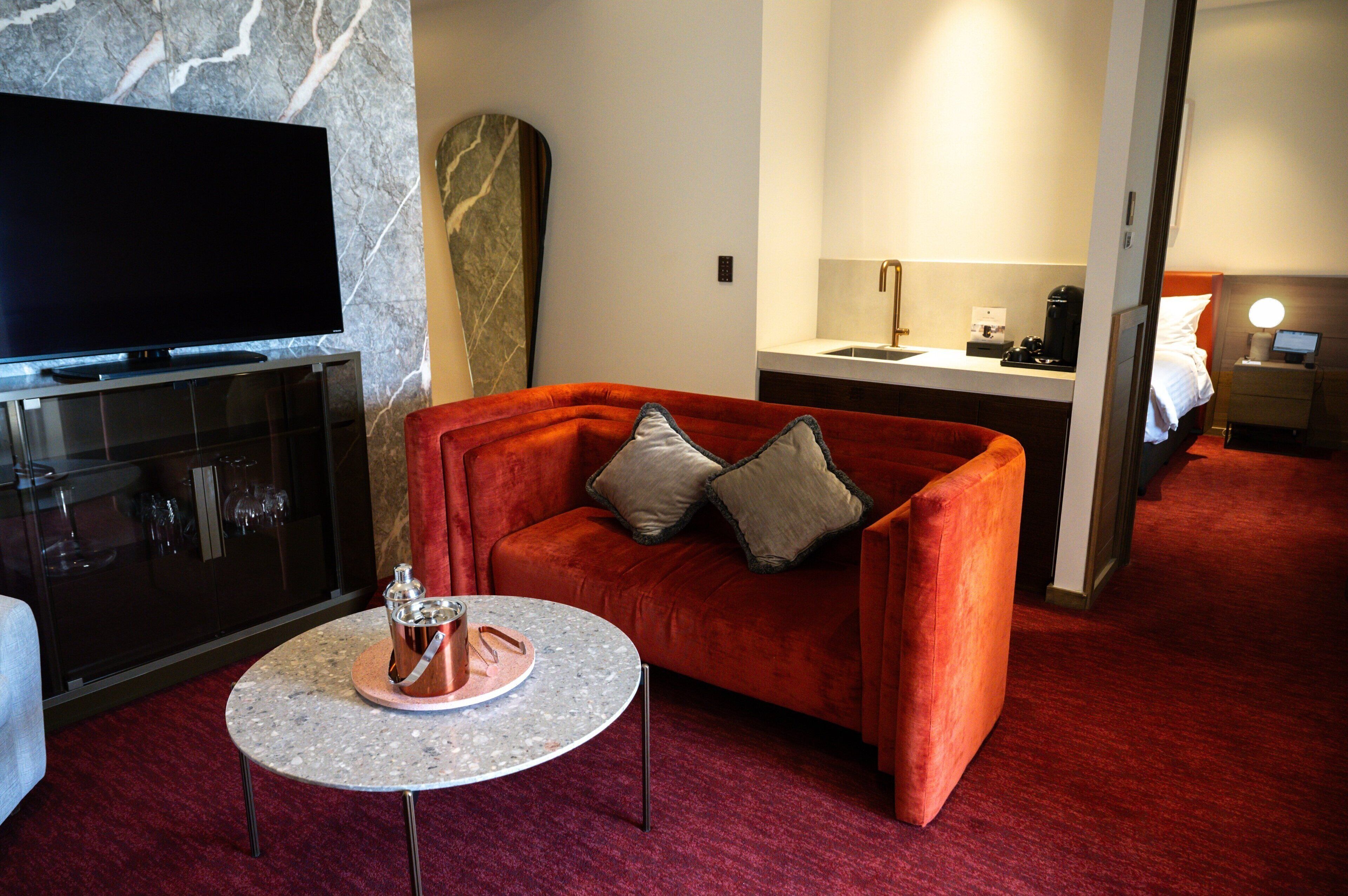 A modern hotel room with a flat-screen TV, a red velvet sofa, a small round table with a tray, and a marble-patterned wall.