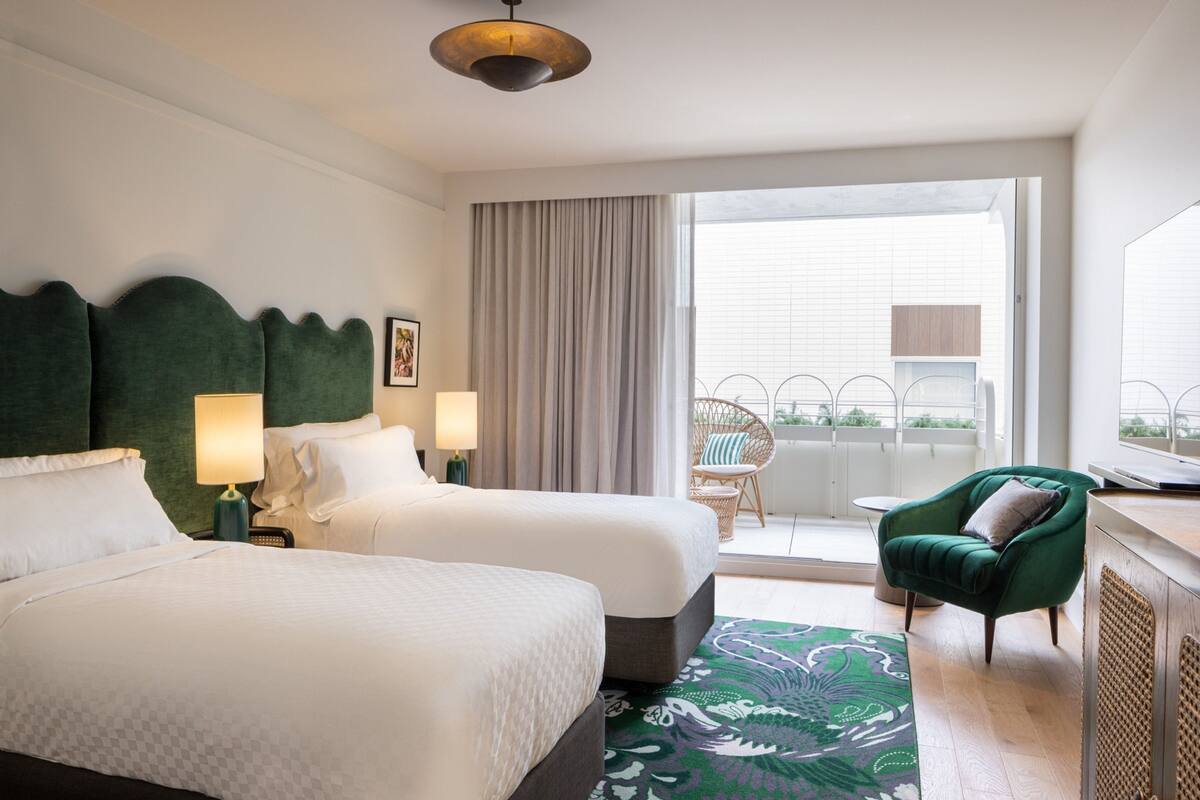 Een hotelkamer met twee bedden, een groene fauteuil, een raam met gordijnen en een balkon met een rieten stoel.