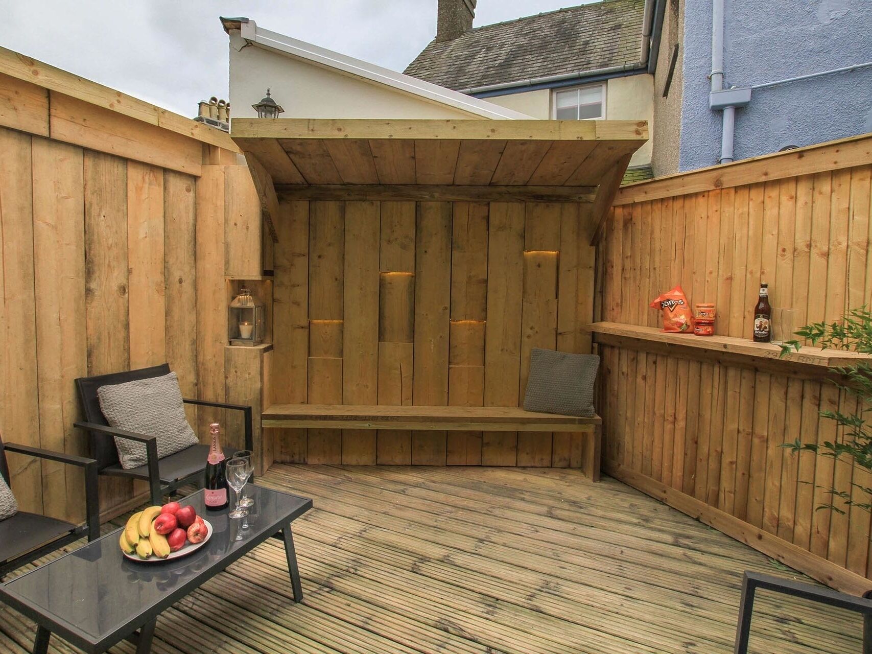 Una terraza de madera con un banco, una mesa con frutas y vasos, y un estante con aperitivos y una botella.