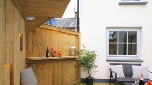 A wooden deck with a bench, a table with wine glasses and apples, a potted plant, and a window with a view of the sky. - Little Bakehouse (Criccieth)
