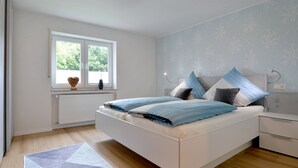A modern bedroom with a large bed, a window with a view of greenery, a radiator, and a bedside table. - Vacation apartment I Kleine - Eifel - Time out (Hambuch)