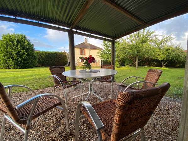 Eine überdachte Terrasse mit einem runden Tisch und Korbstühlen, mit Blick auf eine gepflegte Rasenfläche und ein Haus im Hintergrund.
