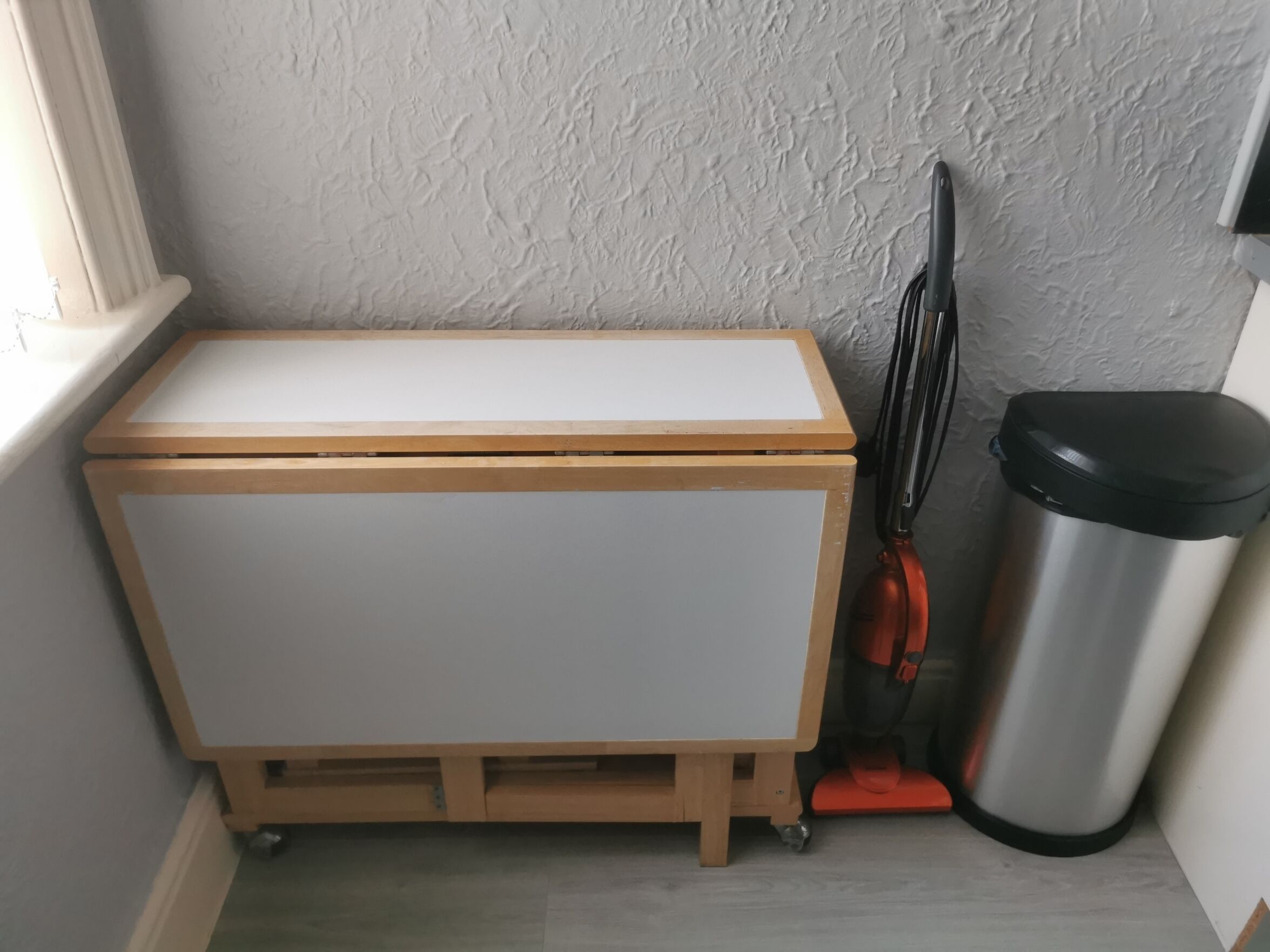 A small wooden cabinet with a white top and drawers, placed next to a trash bin and a window.