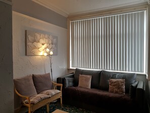 Ein Wohnzimmer mit einem dunkelbraunen Sofa, einem Korbstuhl und einem großen Fenster mit vertikalen Jalousien.