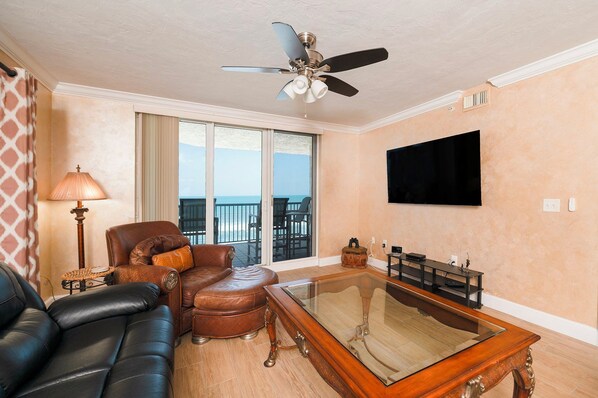 A living room with a leather sofa, a brown leather chair, a glass coffee table, a ceiling fan, a TV, and a balcony with a view of the ocean.