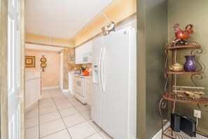 A compact kitchen with white appliances, a microwave, and a toaster. There is a decorative rooster on a shelf and a small birdcage on the floor.