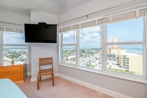A bedroom with a large window, a TV mounted on the wall, a wooden dresser, and a wicker chair. - 10th Floor Heaven at 1001 Towers 10 3/3 Oceanfront (Daytona Beach Shores)