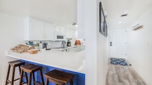 Cocina con gabinetes blancos, microondas y cafetera. Hay taburetes y una encimera azul.