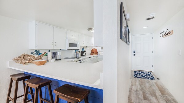 Cocina con gabinetes blancos, microondas y cafetera. Hay taburetes y una encimera azul.