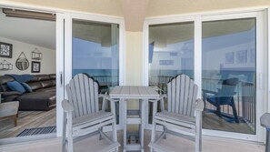 A balcony with a table and two chairs, a view of the ocean, and a living room with a couch and decorative items.