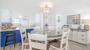 Una cocina moderna con una isla central, armarios blancos y un comedor con mesa de vidrio y sillas blancas.