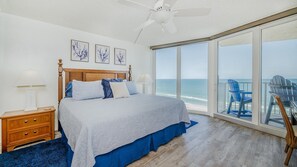 Un dormitorio con una cama grande, ventilador de techo, mesita de noche y vista al océano a través de amplias ventanas.