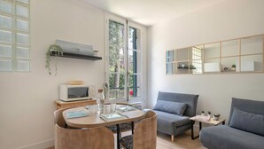 Un salon moderne comprenant un coin repas, des canapés gris, une petite table avec une plante et une grande fenêtre donnant sur des arbres.