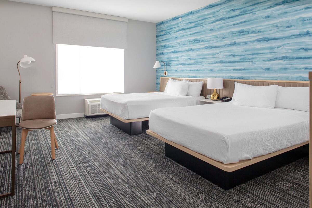 Habitación de hotel con dos camas, un escritorio, una silla, una lámpara y un ventanal grande.