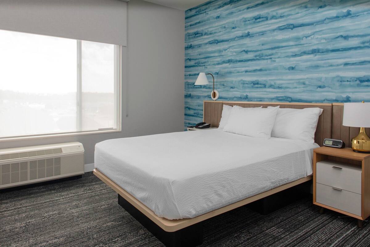 Habitación de hotel con una cama grande, una mesita de noche, una lámpara, un calentador y vistas a la ciudad a través de una ventana.
