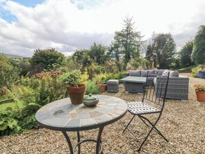 A garden patio with a round table, two potted plants, a wicker sofa, and a metal chair.