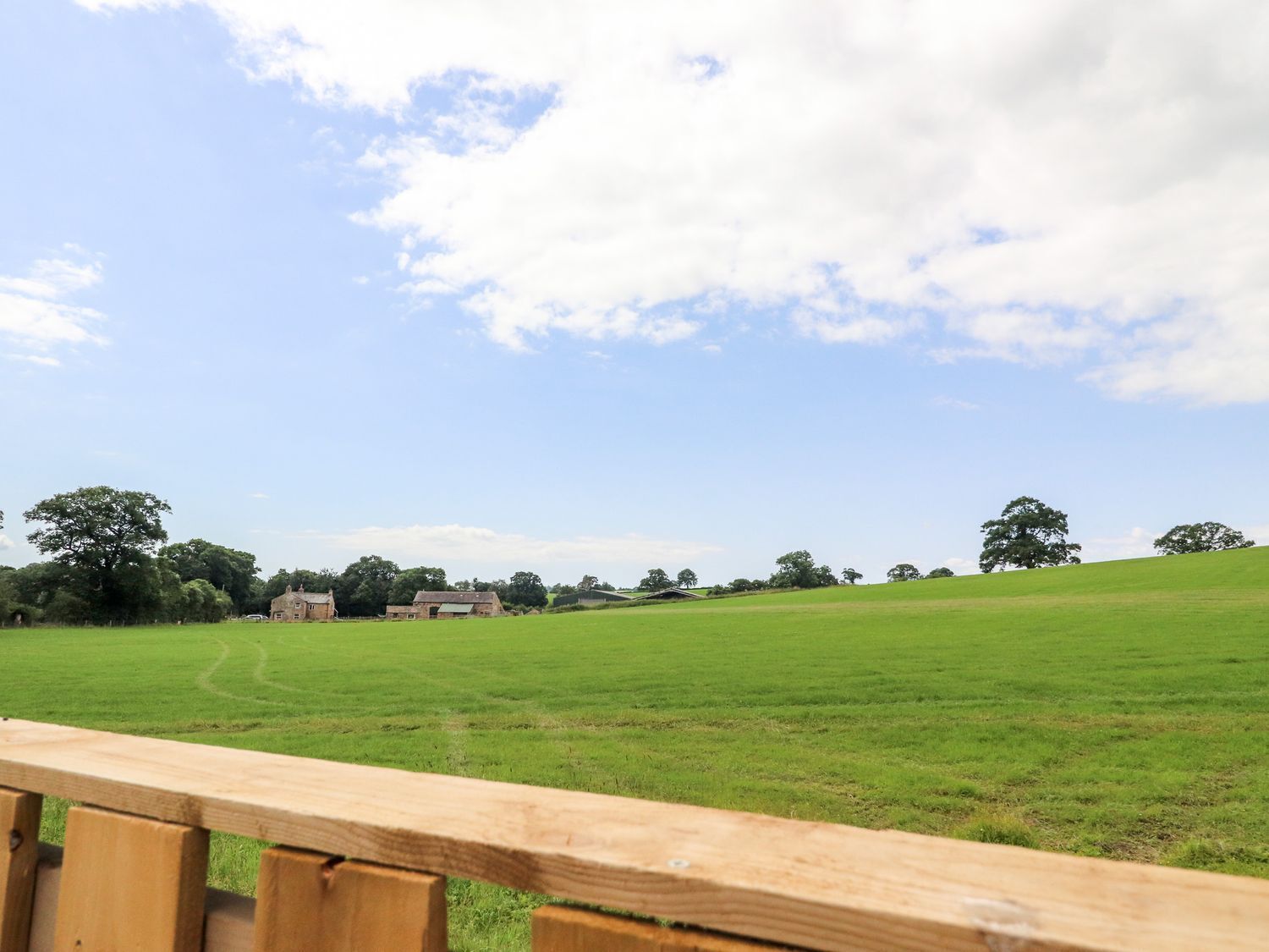 A grassy field with a wooden fence in the foreground and a few buildings in the distance under a partly cloudy sky.
