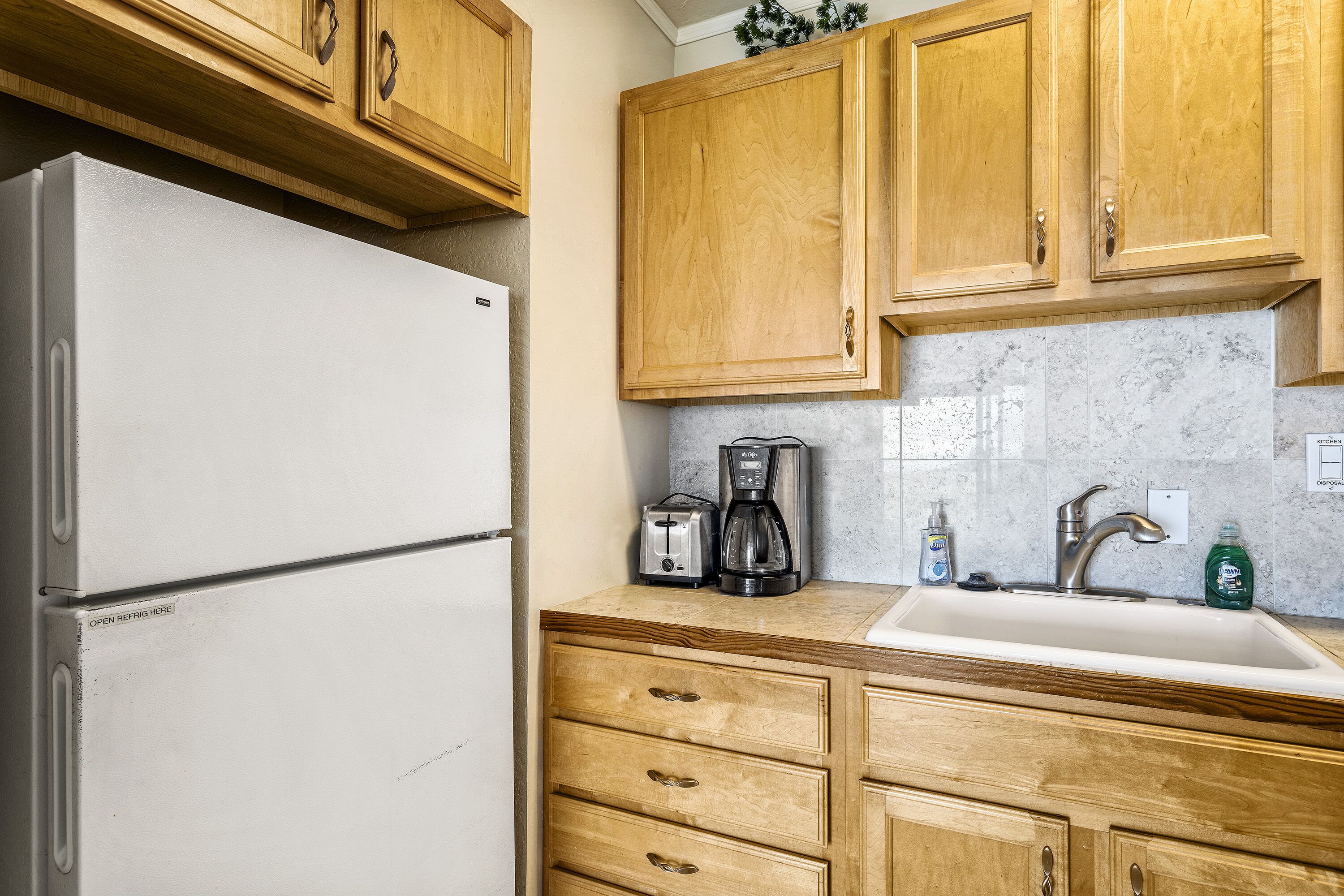 Una cocina con armarios de madera, un refrigerador blanco, una cafetera y un lavamanos.