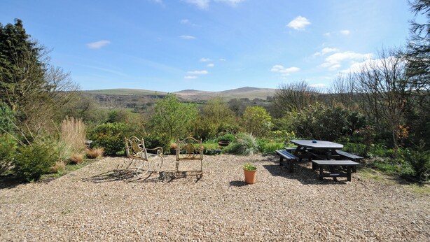 A gravel area with metal chairs and a picnic table, surrounded by greenery and hills in the background. - Tor View @ Fourwinds (Okehampton)