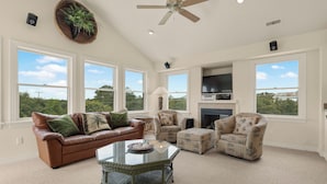 A spacious living room with a brown leather couch, a glass coffee table, wicker chairs, and large windows offering a view of greenery. - The Hidden Gem: Hot Tub, Private Pool, Currituck Club Amenities (Corolla)
