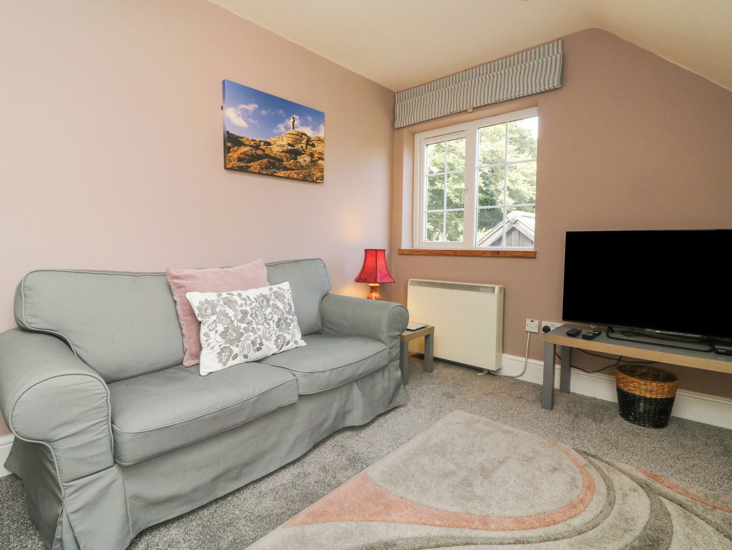 A living room with a grey sofa, a flat-screen TV, a radiator, a window with a view of trees, and a painting on the wall.