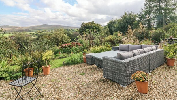 Un patio con muebles de mimbre y plantas en macetas, con vistas a un jardín exuberante y colinas lejanas.