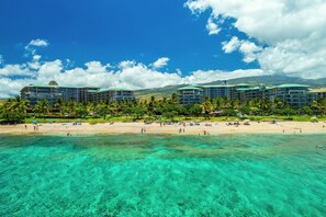 Un hotel frente a la playa con varias construcciones, aguas turquesas cristalinas y una playa arenosa con sombrillas y gente.