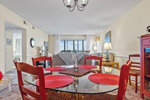 A dining area with a glass table surrounded by red chairs, a chandelier, and a view of the outdoors through large windows. - Point Prospect Shores- Ocean Front Condo (Pine Knoll Shores)