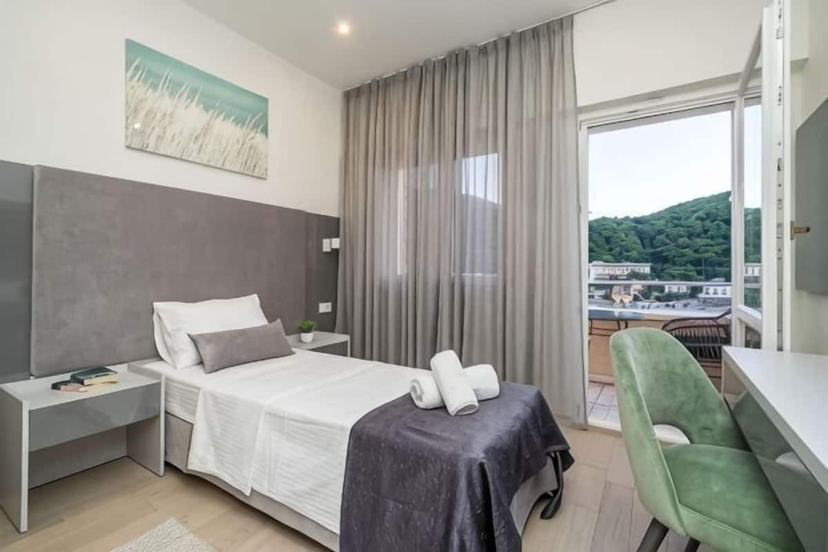 A modern hotel room with a bed, a desk, a chair, a view of the city, and a balcony.