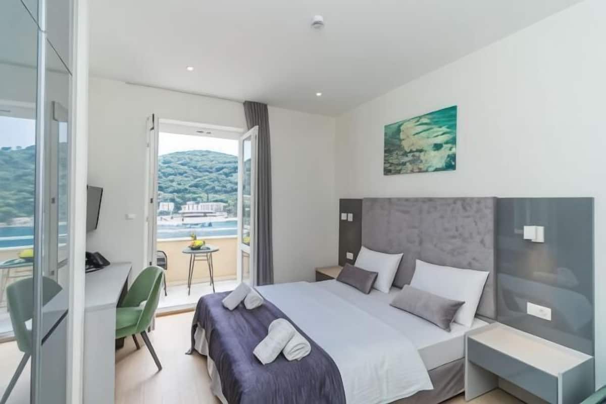 A hotel room with a bed, a desk, a chair, a balcony with a view of the sea, and a painting on the wall.
