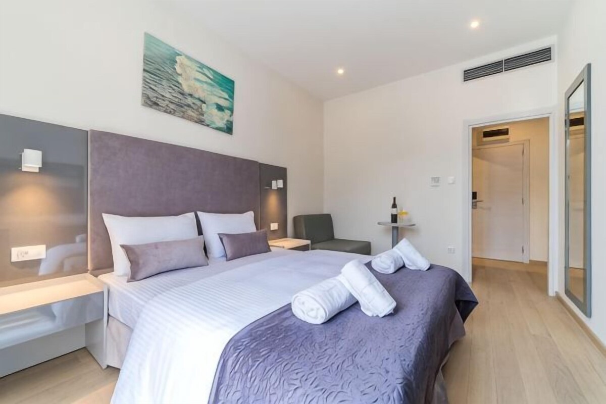 A modern hotel room with a large bed, bedside tables, a desk, and a painting on the wall.