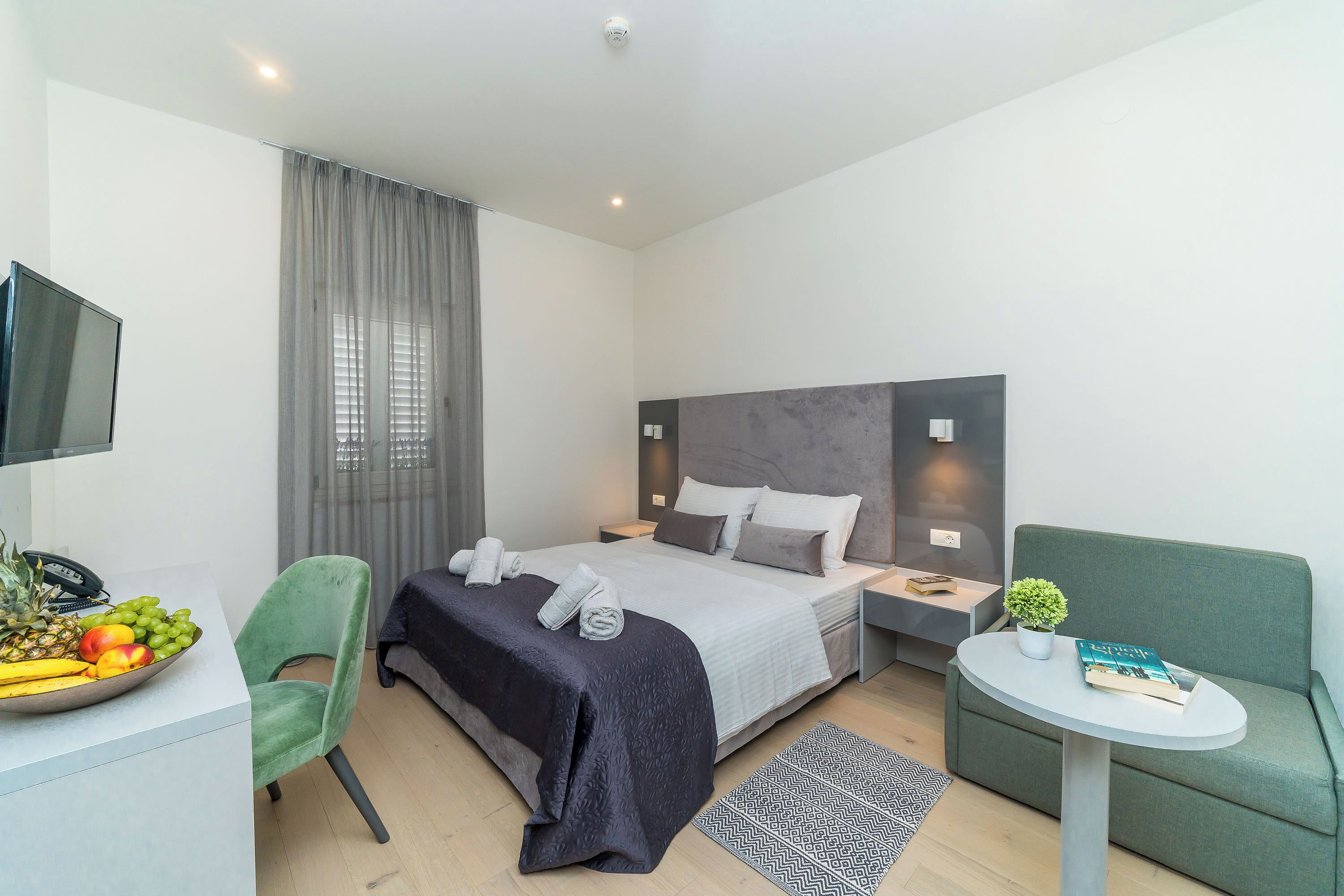Une chambre d’hôtel moderne avec un lit, un bureau agrémenté d’un panier de fruits, une chaise verte, une petite table avec un livre et une plante, et un canapé gris.