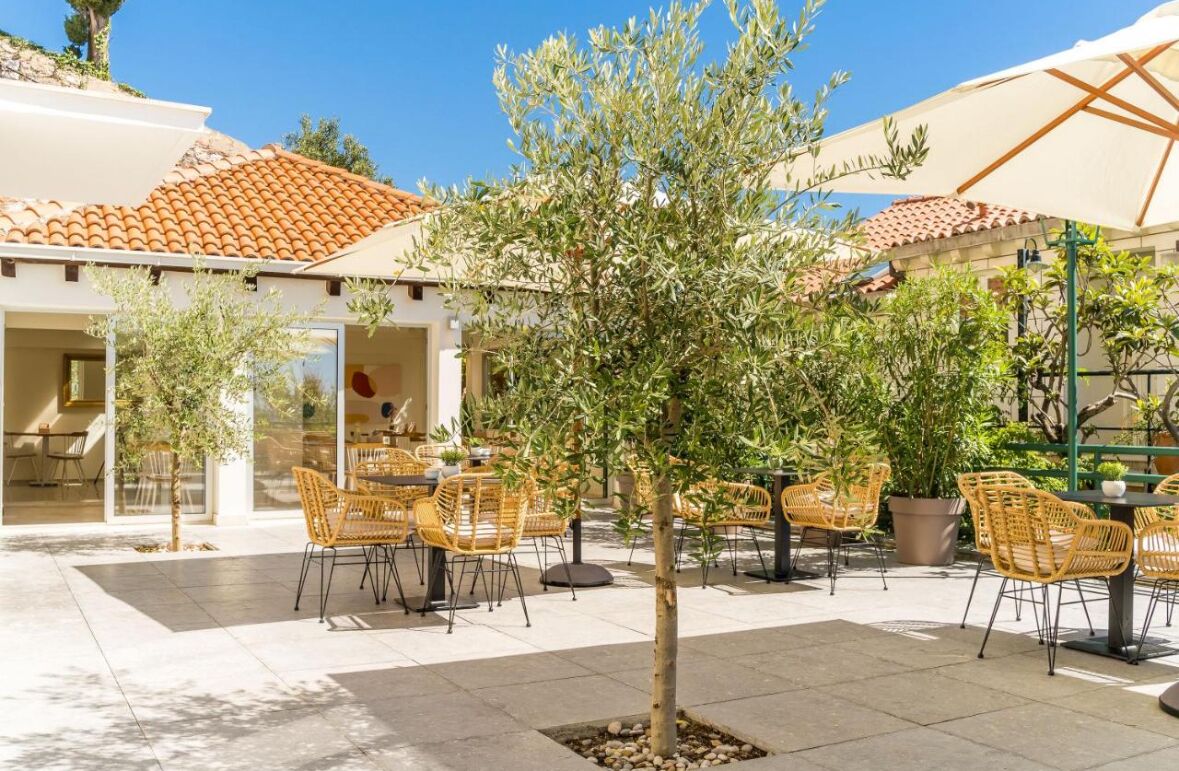 An outdoor patio area with wicker chairs and tables, surrounded by potted plants and a tree.
