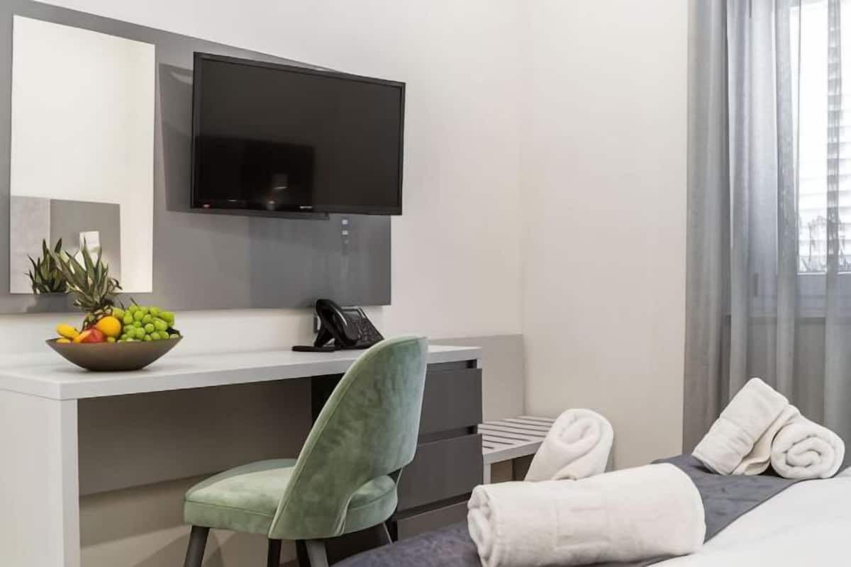 A hotel room with a bed, a desk with a chair, a television, a bowl of fruit, and a window with curtains.