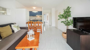 Un salon moderne avec un canapé, une table en bois, un téléviseur et un coin repas comprenant des chaises, avec une cuisine en arrière-plan.