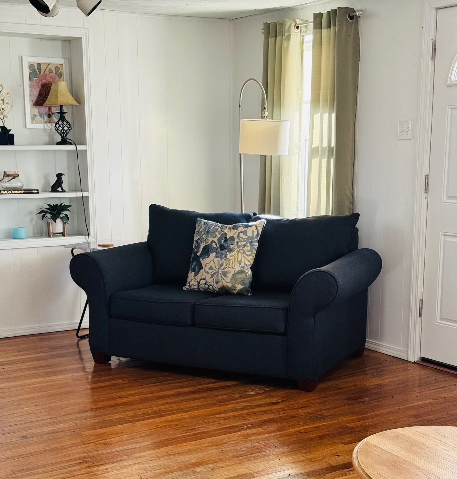 A dark blue sofa with floral cushions in a room with white walls, a wooden floor, a lamp, and a window with curtains.