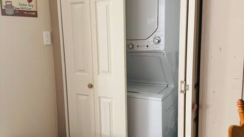 Uma lavanderia com máquina de lavar e secadora brancas, uma porta branca e uma lixeira.