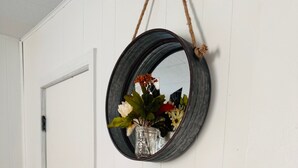 Een witte dressoir met drie lades, een spiegel met een touwhandvat, en een vaas met bloemen aan de muur.
