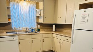 Een keuken met houten vloeren, beige kastjes, een witte koelkast en een raam met een blauw gordijn.