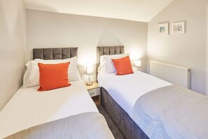 Une chambre d’hôtel avec deux lits, chacun doté d’une tête de lit, de draps blancs et d’un oreiller orange. On y trouve des tables de chevet avec des lampes, un radiateur et des tableaux encadrés accrochés au mur.
