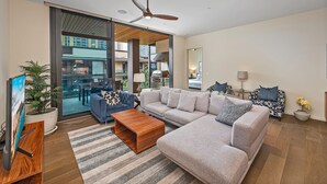 Ein modernes Wohnzimmer mit einem großen Ecksofa, einem Couchtisch und einem Fernseher auf einem Holzständer. Es gibt einen Deckenventilator, ein großes Fenster und einen Blick ins Freie.