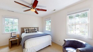A bedroom with a bed, a chair, a nightstand, a ceiling fan, and two windows.