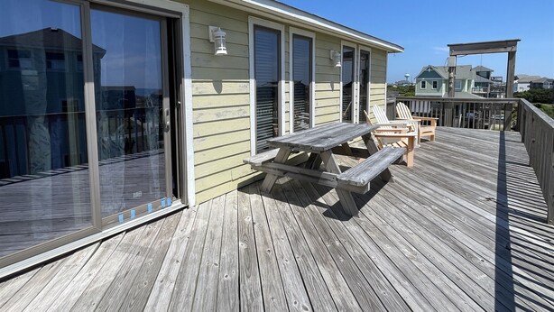 A wooden deck with a picnic table and chairs, a glass door, and a building with shutters. - Southern Star 5 Bedroom Cottage by RedAwning (Hatteras)